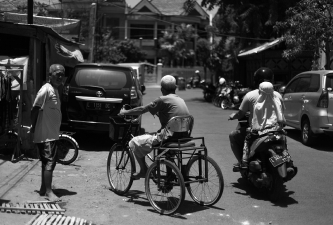 Three elderly men interacting on a sunlit urban street, one riding a tricycle, another on a motorbike, and the third standing barefoot near a building, with parked cars and houses in the background, creating a lively and candid atmosphere